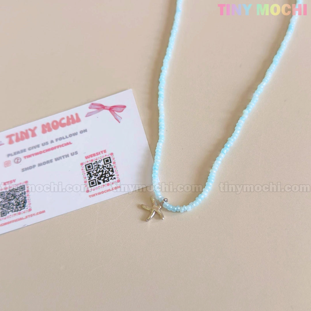 The Silver Starfish Choker Necklace by Tiny Mochi features light blue beads and a dainty starfish charm, perfect oceancore jewelry for girls or teens—shown with their pastel business card adorned with QR codes on a beige background.