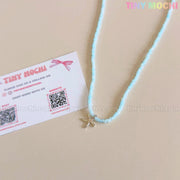 The Silver Starfish Choker Necklace by Tiny Mochi features light blue beads and a dainty starfish charm, perfect oceancore jewelry for girls or teens—shown with their pastel business card adorned with QR codes on a beige background.