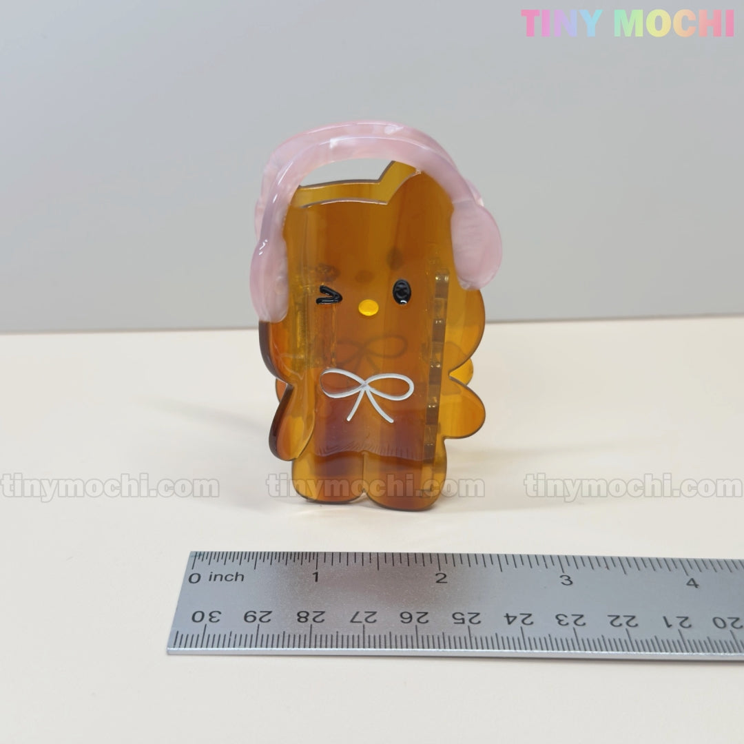 A cute translucent brown bear hair claw clip with pink headphones from Tiny Mochi stands next to a metal ruler. This kawaii accessory matches perfectly with any pastel lace ribbon keychain. The Tiny Mochi logo appears in pastel at the top right.