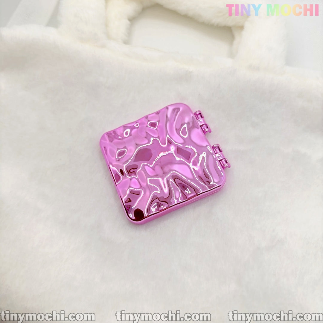 A shiny, metallic pink square double-sided compact mirror with a wavy texture from Tiny Mochi rests on a fuzzy white surface. This portable, Y2K aesthetic makeup accessory features “TINY MOCHI” in pastel at the top right corner.
