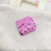 A shiny, metallic pink square double-sided compact mirror with a wavy texture from Tiny Mochi rests on a fuzzy white surface. This portable, Y2K aesthetic makeup accessory features “TINY MOCHI” in pastel at the top right corner.