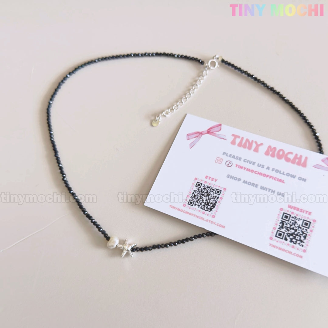 The Sterling Silver Starfish Necklace by Tiny Mochi—a handmade 925 silver charm on a black beaded pearl choker—rests next to a Tiny Mochi card displaying social media and website info on a light pink background.