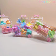 Colorful Kids Hair Clips from Tiny Mochi, in shapes like butterflies, hearts, and flowers, are packed in small transparent mini travel cases scattered on a light surface. The brand name Tiny Mochi appears in the top right corner.