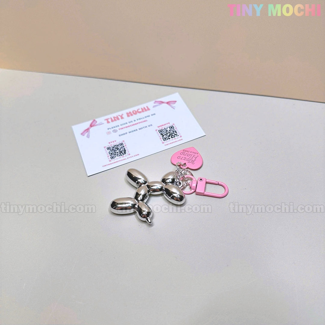 The Tiny Mochi Balloon Dog Keychain Chrome Silver Bag Charm, featuring a cute pink heart tag, shines on a light surface next to a business card. The pastel Tiny Mochi logo appears at the top right.
