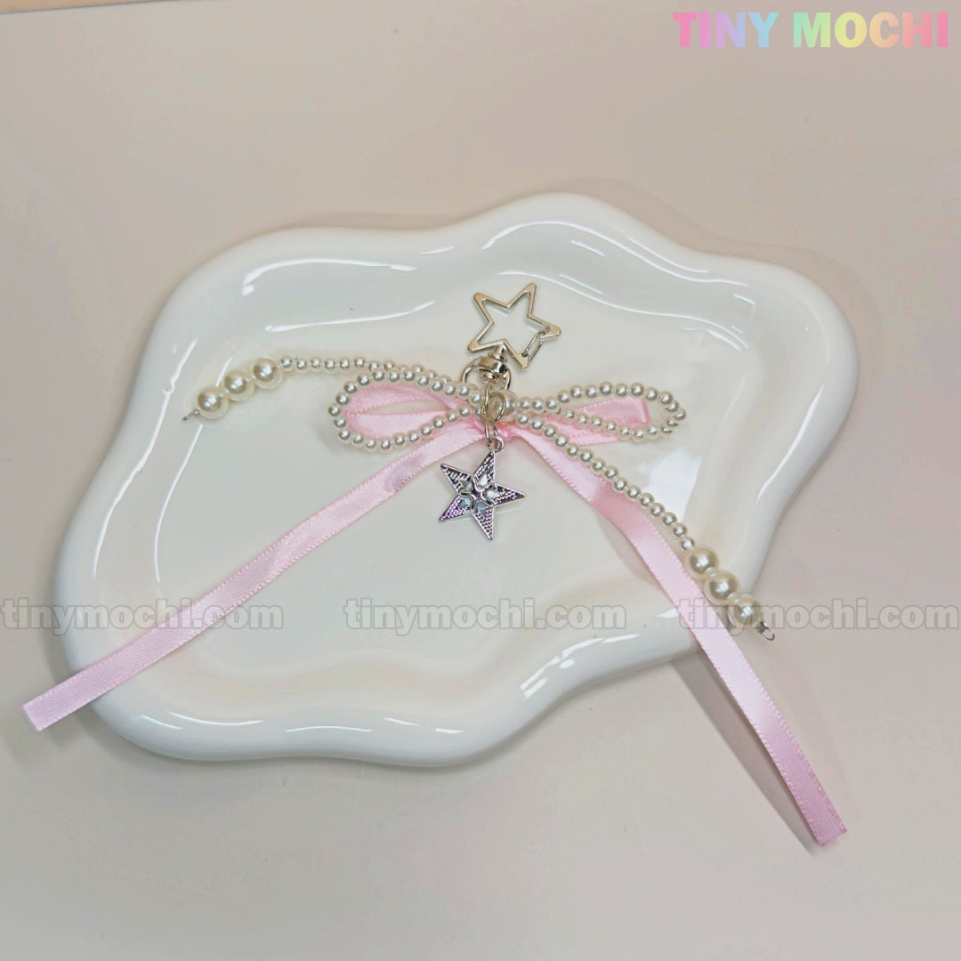 The Pink Star Bow Charm Keychain by Tiny Mochi, with Y2K ribbon, pearls, and silver star details, is shown on a white wavy dish—a kawaii coquette bag or phone accessory. TINYMICHI.COM is subtly visible.