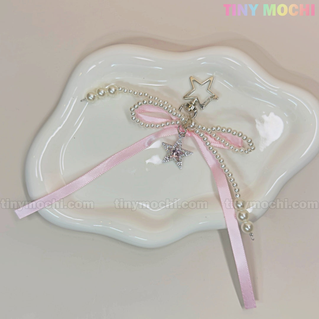 A Pink Star Bow Charm Keychain with pearls and a pink ribbon from Tiny Mochi is displayed on a wavy white ceramic dish. A pastel TINY MOCHI watermark appears in the upper right corner.