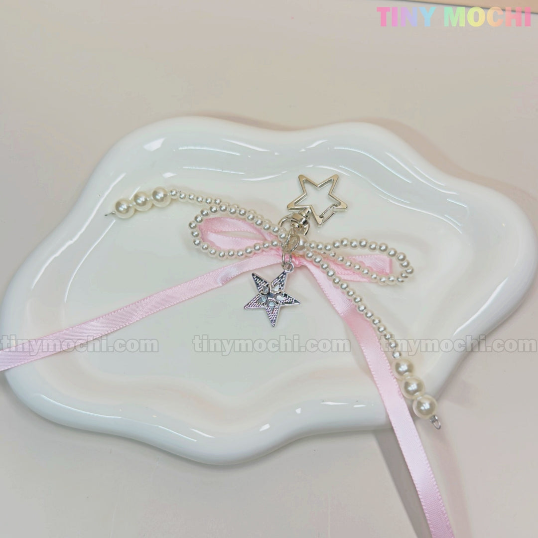 The Pink Star Bow Charm Keychain by Tiny Mochi features kawaii Y2K ribbon and pearl accents. Displayed on a white cloud-shaped dish with a pastel background, its the perfect coquette bag accessory or phone charm gift.