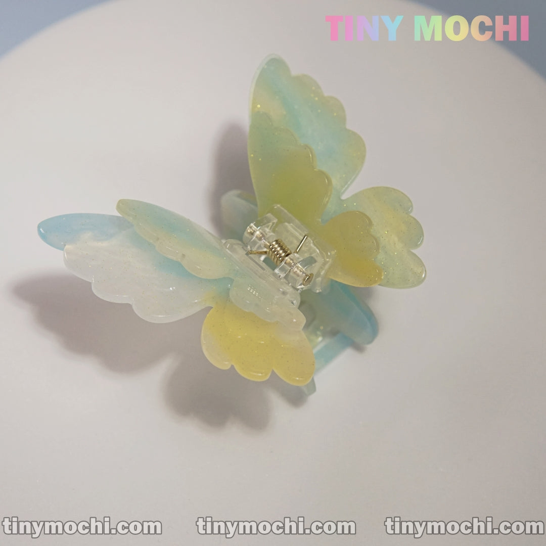 A pastel-colored Butterfly Hair Claw Clip by Tiny Mochi, featuring UV changing colors and blue, yellow, and translucent wings, is displayed on a white surface with Tiny Mochi in the top right and tinymochi.com along the bottom.