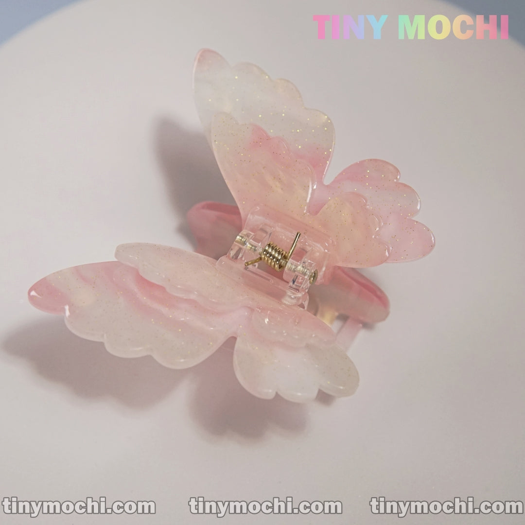 A pastel pink and white Butterfly Hair Claw Clip by Tiny Mochi, featuring UV color-changing acetate with a subtle glittery finish, is shown on a light surface with rainbow TINY MOCHI lettering and tinymochi.com below.