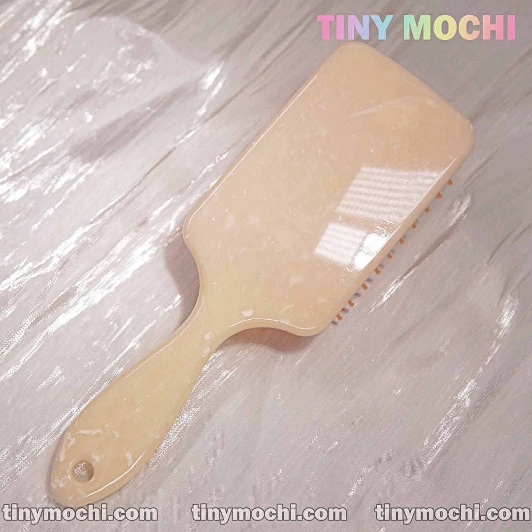 A glossy beige Marble Pattern Detangling Brush—Nordic Style Paddle Hair Brush for Dry and Wet Hair by Tiny Mochi—rests on a textured surface. Tiny Mochi appears in pastels at the top right, with tinymochi.com at the bottom.