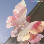 A close-up of a pastel pink and white Tiny Mochi Butterfly Hair Claw Clip, UV Changing Color Hair Clip with a marbled pattern, crafted from durable acetate, shown on a reflective surface. TINY MOCHI appears in the top right and bottom edges.