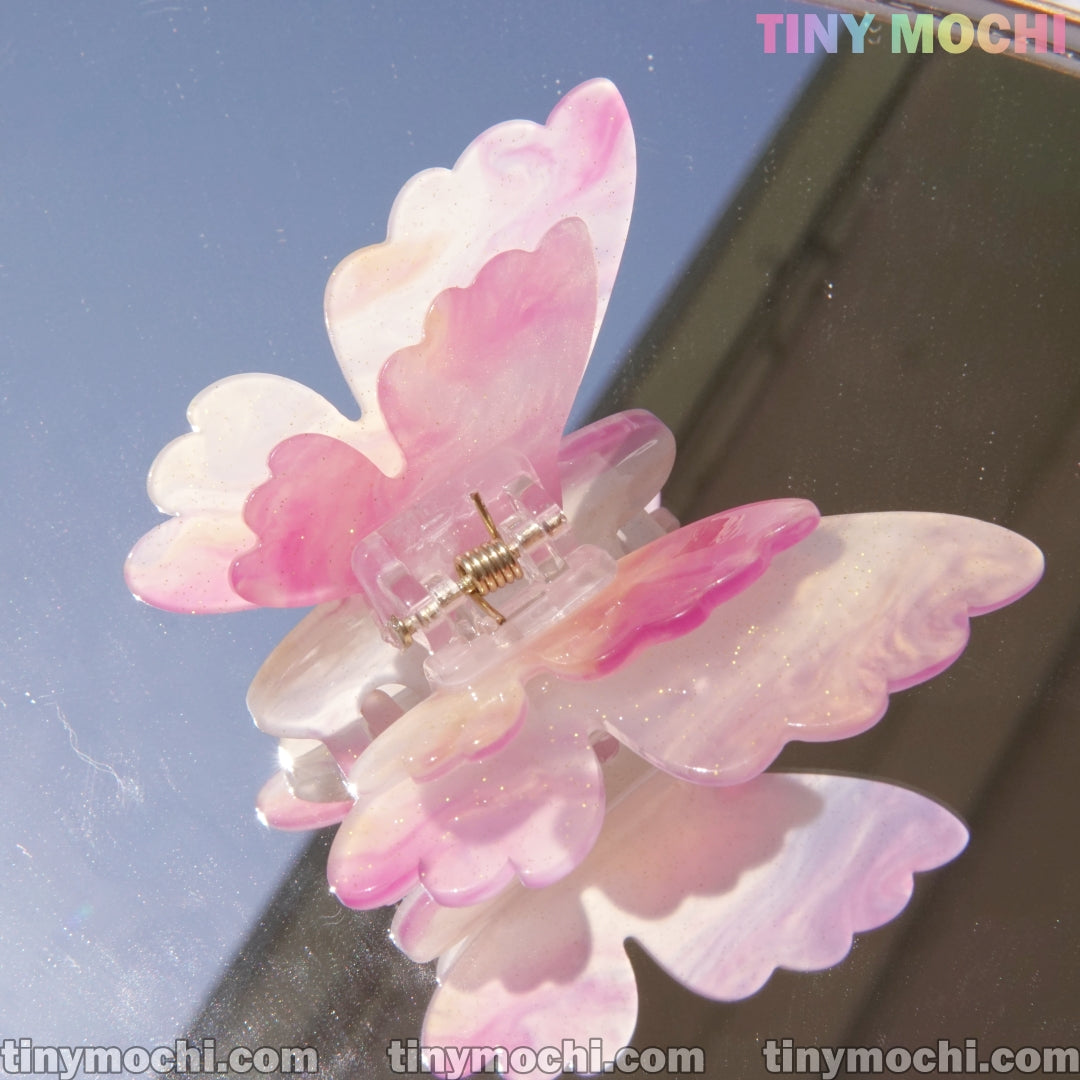 A glossy pink and white Butterfly Hair Claw Clip from Tiny Mochi rests on a reflective surface. This shimmery UV changing color hair accessory is crafted from durable acetate and displays the Tiny Mochi logo in the corner.