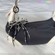 A black and white handbag features Tiny Mochis Elegant Sparkling Bow Bag Charm, adorned with a crystal bowknot and clear heart pendant, displayed on a white surface with Tiny Mochi branding in the corners.