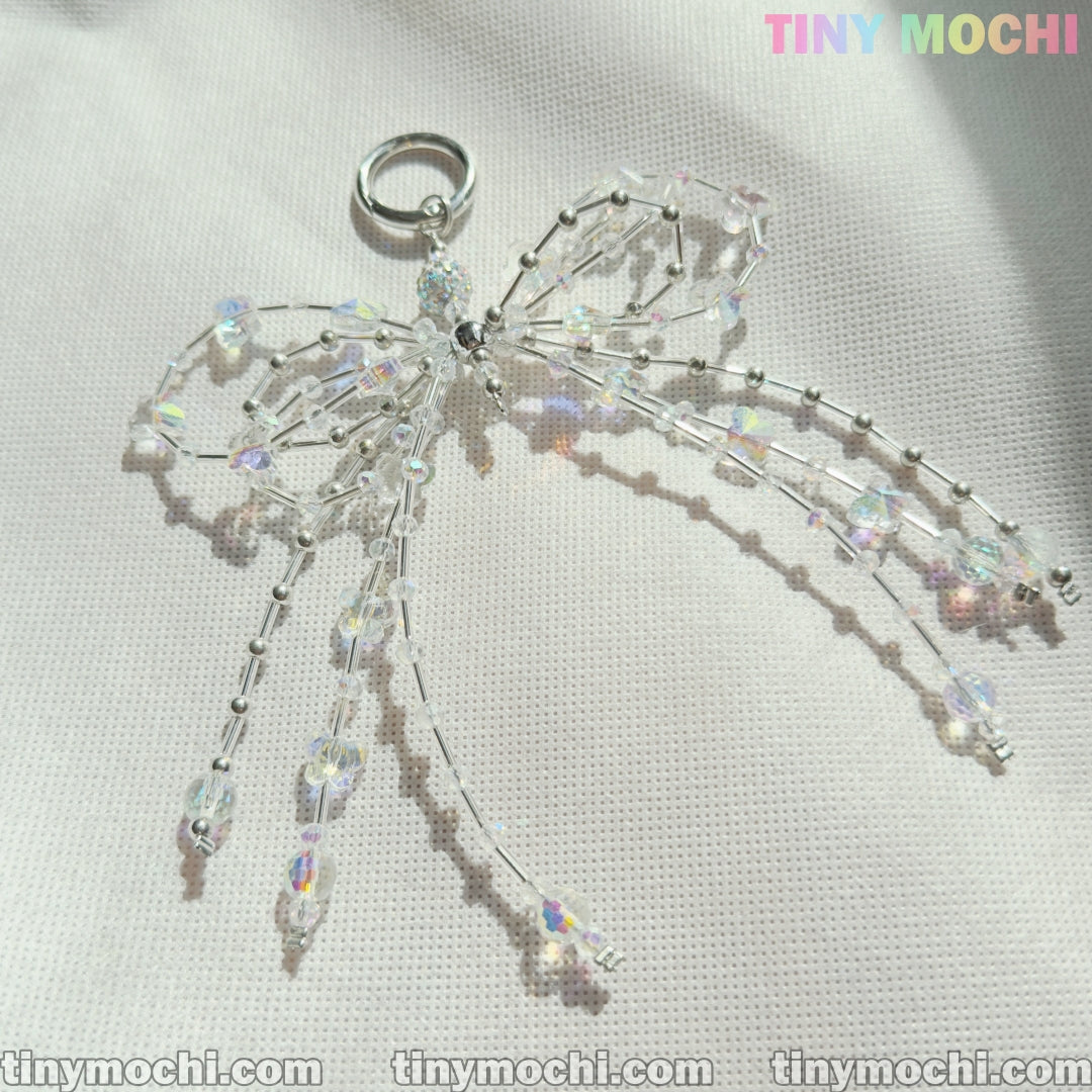 The Tiny Mochi Crystal Bow Pearl Keychain, crafted with clear iridescent beads and silver wire, sparkles on a light textured surface—an aesthetic accessory perfect for bags, keys, and more.