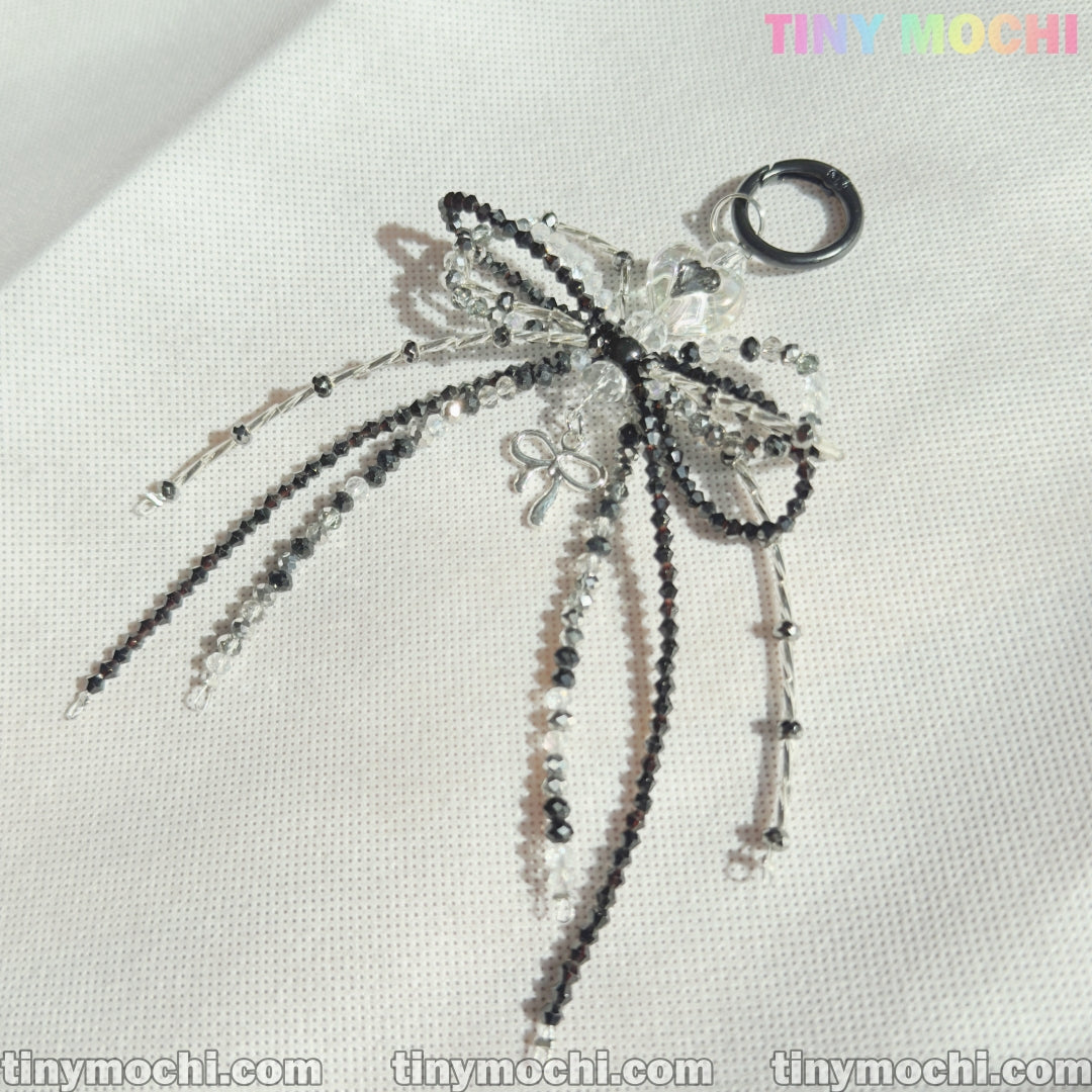 The Elegant Sparkling Bow Bag Charm by Tiny Mochi features a handcrafted beaded bow with black and clear strands, shown on a white textured surface. The image includes the Tiny Mochi watermark in the corners.