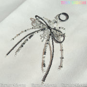 The Elegant Sparkling Bow Bag Charm by Tiny Mochi features a handcrafted beaded bow with black and clear strands, shown on a white textured surface. The image includes the Tiny Mochi watermark in the corners.