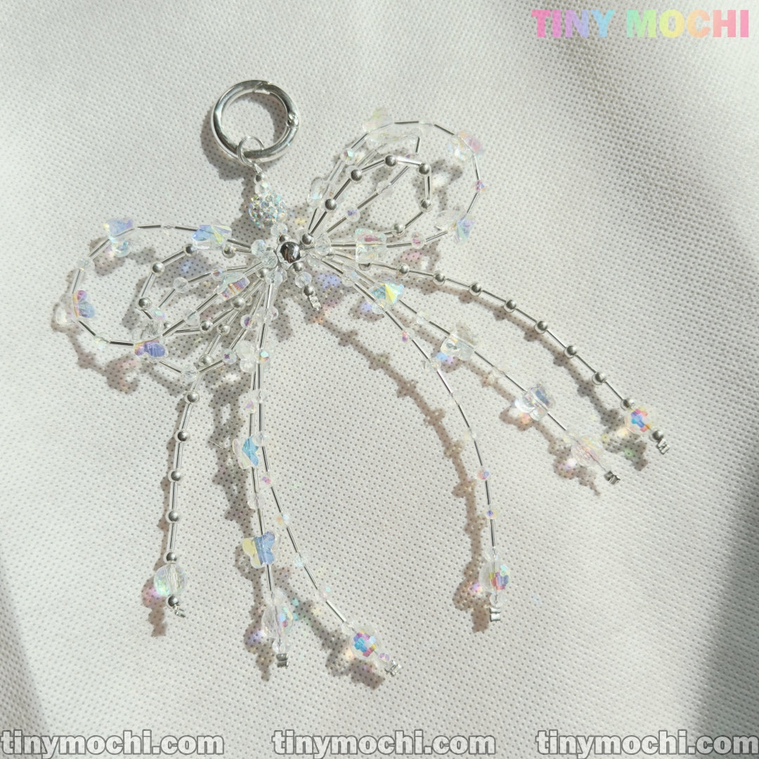 The Crystal Bow Pearl Keychain by Tiny Mochi, featuring iridescent and clear beads in a delicate bow design, is shown on a textured white surface. TINYMochi.com appears in the corners and TINYMOCHI at the top right.
