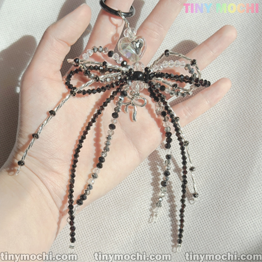 A hand holds the Elegant Sparkling Bow Bag Charm by Tiny Mochi—a handcrafted black and silver beaded accessory with long strands, shimmering in sunlight. Tiny Mochi appears in colorful text at the top right.