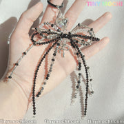 A hand holds the Elegant Sparkling Bow Bag Charm by Tiny Mochi—a handcrafted black and silver beaded accessory with long strands, shimmering in sunlight. Tiny Mochi appears in colorful text at the top right.