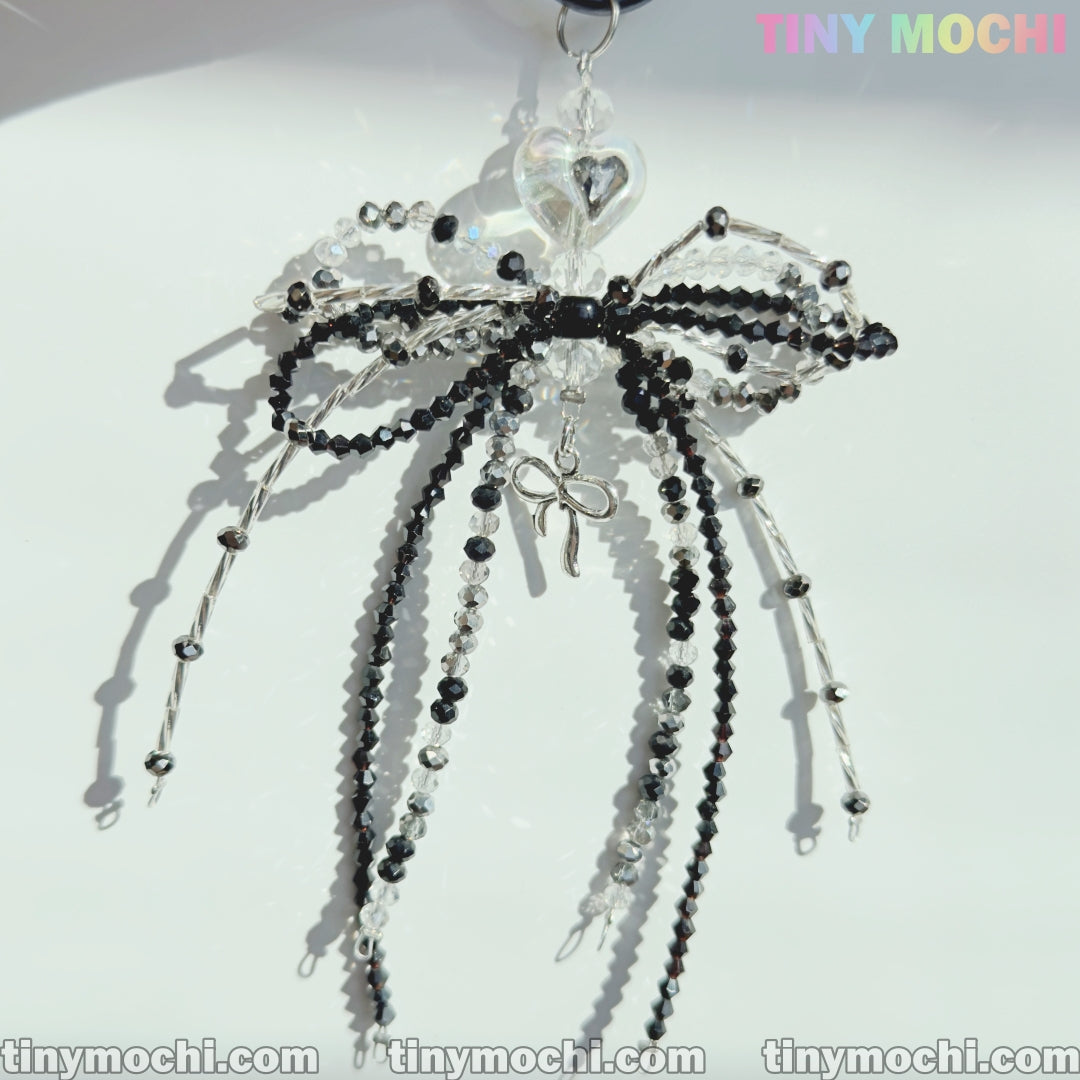 The Elegant Sparkling Bow Bag Charm by Tiny Mochi features a handcrafted crystal bowknot with black and clear beads, dangling strands, and a silver ribbon on a white background with Tiny Mochi in pastel colors at the top right.