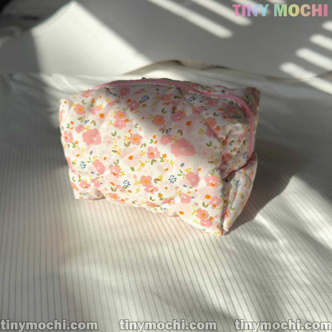 A Tiny Mochi Floral Quilted Cosmetic Bag (6.7 x 4.5 x 3.9) sits on a white, striped fabric in sunlight—perfect as a beauty case or travel pouch. TINY MOCHI appears at the top right and tinymochi.com along the bottom edge.