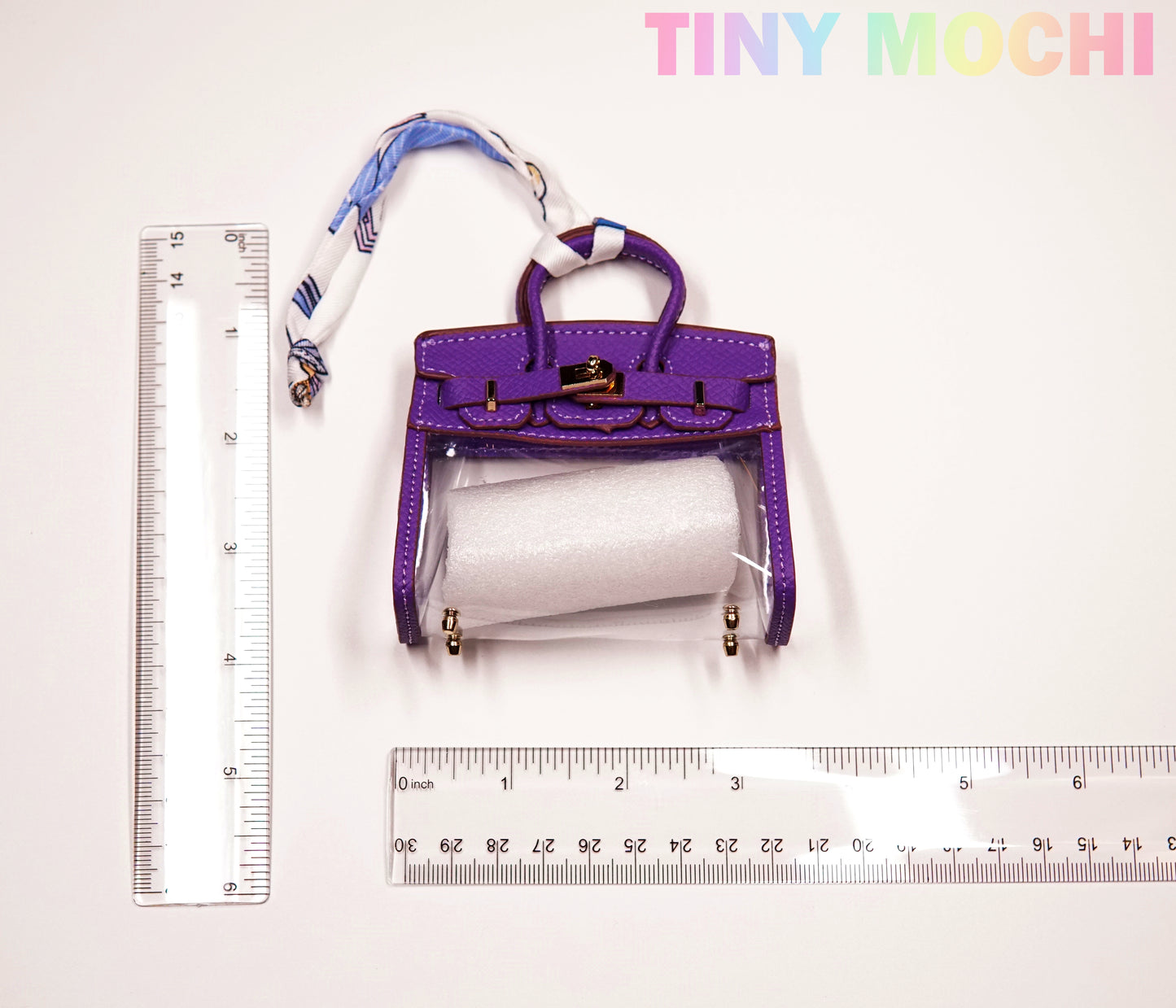 A small Tote-Shaped Transparent Key Pouch from Tiny Mochi, featuring a transparent front and roll of paper inside, is placed between two rulers. The brand name Tiny Mochi appears in colorful letters at the top right.