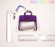A small Tote-Shaped Transparent Key Pouch from Tiny Mochi, featuring a transparent front and roll of paper inside, is placed between two rulers. The brand name Tiny Mochi appears in colorful letters at the top right.
