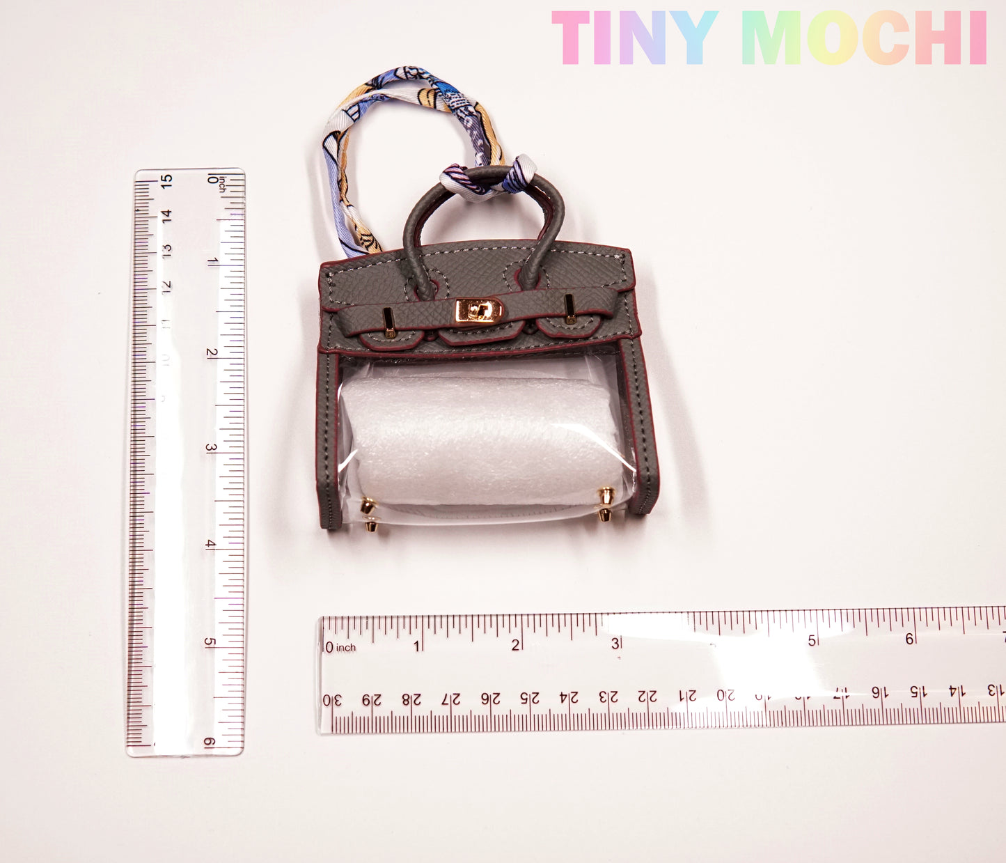 The Tote-Shaped Transparent Key Pouch by Tiny Mochi, a stylish mini bag key holder with gray trim, is shown between two rulers for scale. TINY MOCHI appears in pastel colors at the top right.