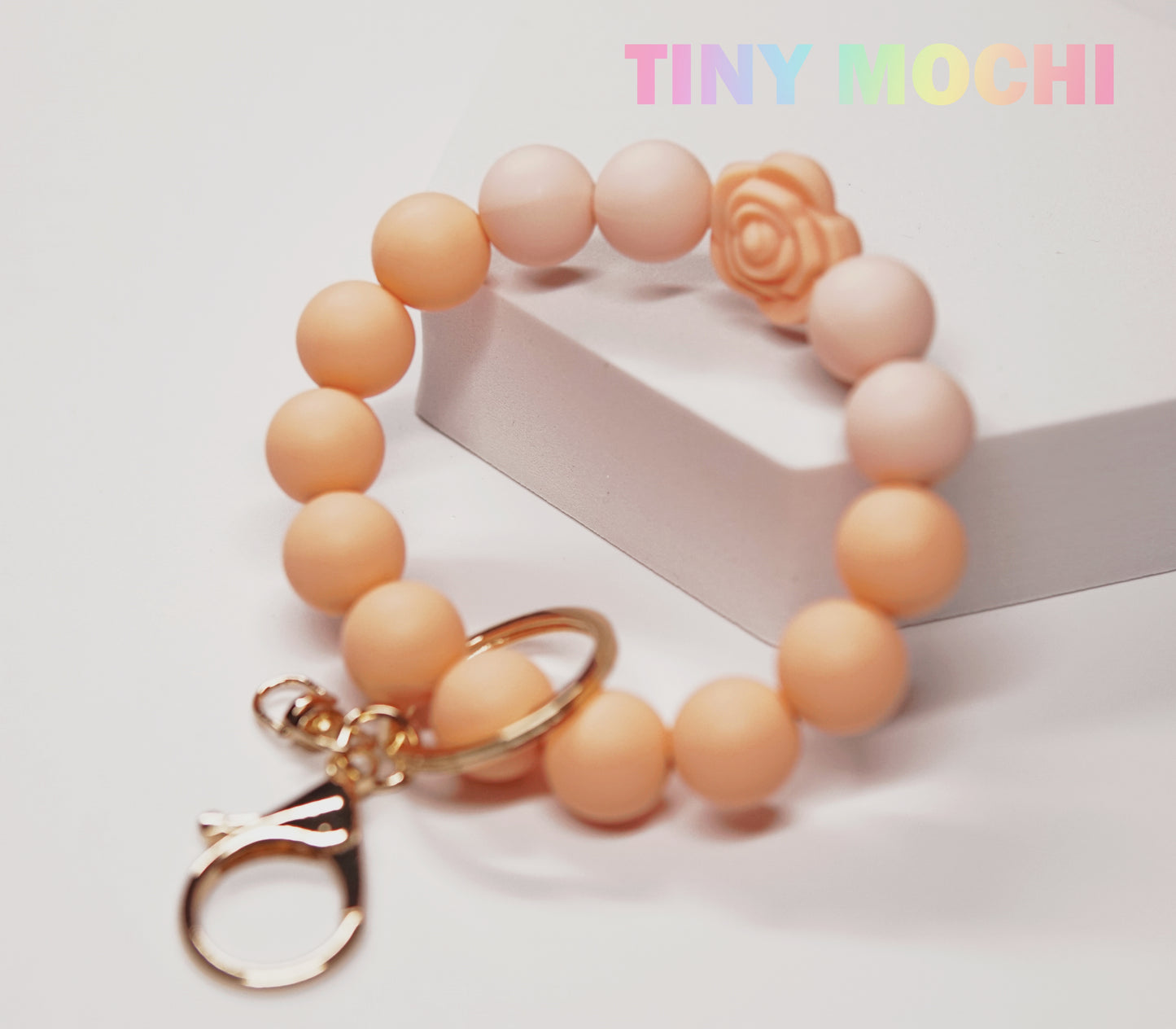 The Adorable Flower Keychain Bracelet by Tiny Mochi features peach and light pink food-grade silicone beads, a rose-shaped bead, and a gold clasp and keyring. TINY MOCHI is displayed in pastel colors at the top right.