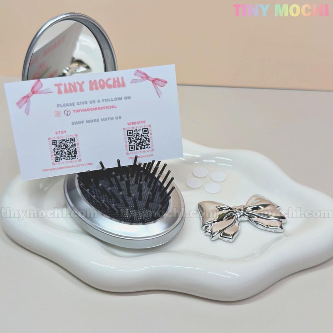 The Tiny Mochi Oval Silver Folding Travel Mirror Comb—a compact pocket-sized brush with built-in mirror—sits with a clip, cushion hairbrush, and business card featuring QR codes on a wavy white tray with three small round stickers.