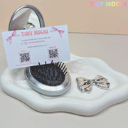 The Tiny Mochi Oval Silver Folding Travel Mirror Comb—a compact pocket-sized brush with built-in mirror—sits with a clip, cushion hairbrush, and business card featuring QR codes on a wavy white tray with three small round stickers.