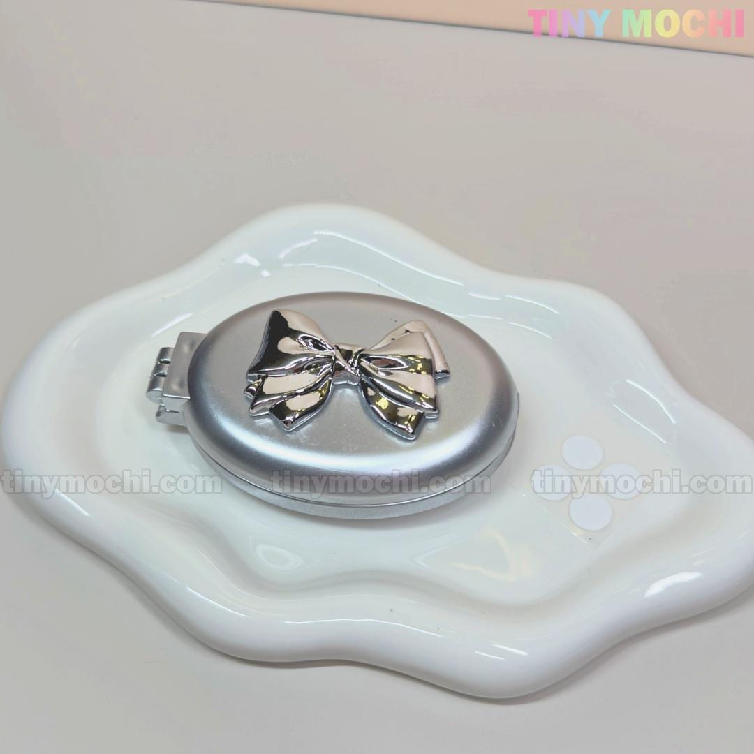 The Tiny Mochi Oval Silver Folding Travel Mirror Comb, a compact pocket-sized brush with built-in mirror for easy on-the-go styling and detangling, rests on a stylish white dish—where tiny mochi is subtly seen in the background.