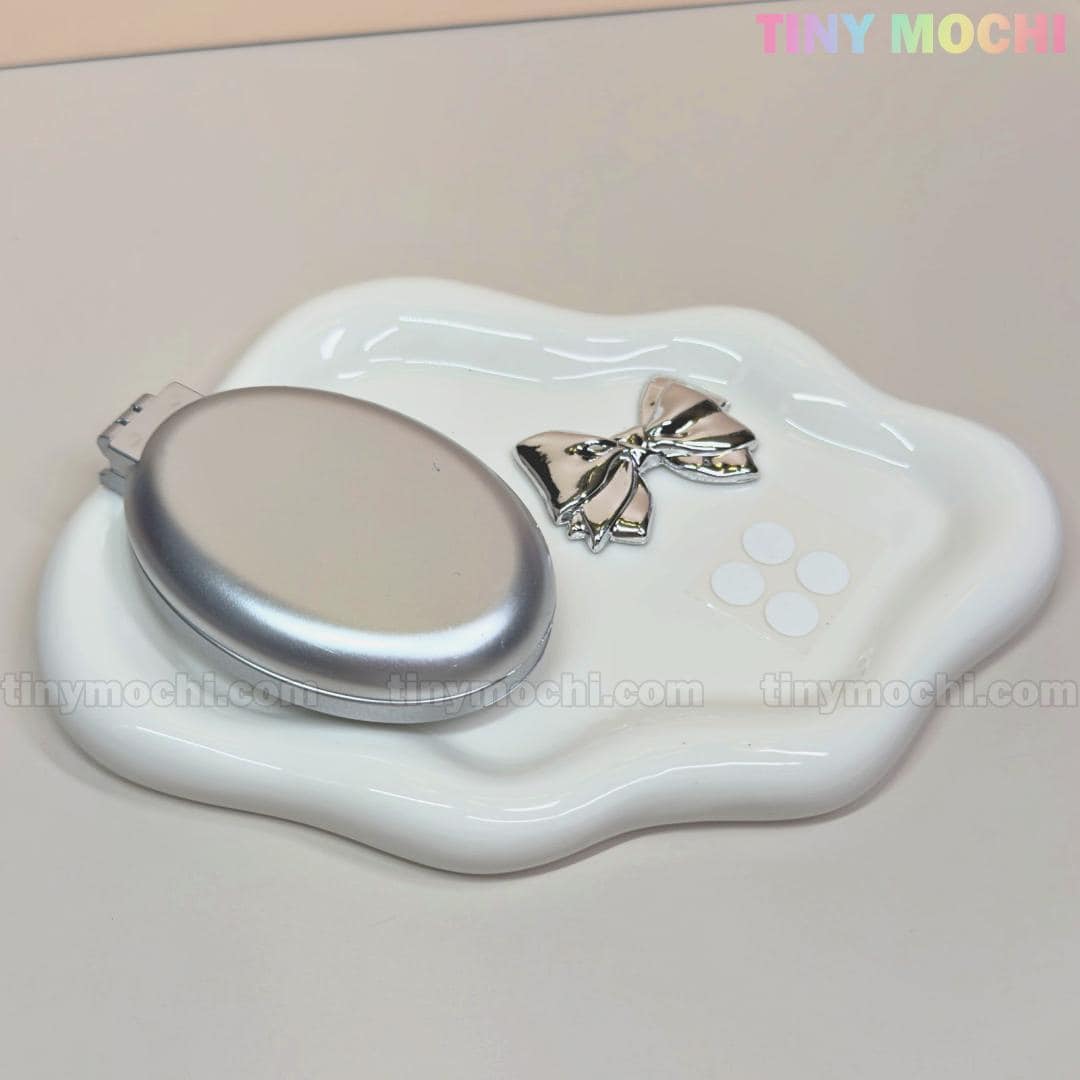 A white dish holds Tiny Mochis Oval Silver Folding Travel Mirror Comb—a compact, pocket-sized hair brush with built-in mirror for easy on-the-go styling and detangling. Pastel TINY MOCHI text appears in the top right corner.