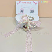 A round white box topped with a pink lace and pearl bow plus the Kawaii Lace Bow Keychain with pearl beads & silver heart charm from Tiny Mochi—ideal for coquette lovers. Behind it is a Tiny Mochi business card with social info and QR codes.
