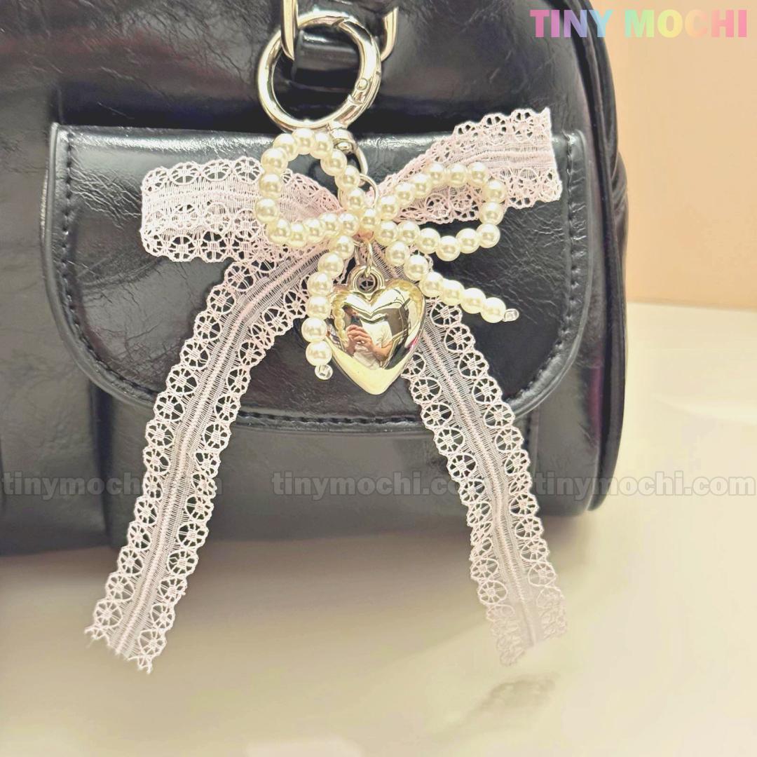 A black bag is adorned with the Kawaii Lace Bow Keychain by Tiny Mochi—featuring a lacy pink bow, pearl beads, and a shiny silver heart charm. The softly blurred background displays the Tiny Mochi logo in the top right corner.