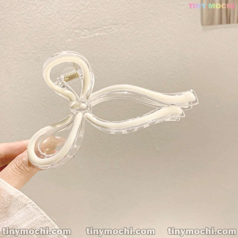 A hand holds the Tiny Mochi Elegant Bow Hair Clip—an adorable, handmade hair accessory crafted from durable plastic with white accents. “tinymochi.com” is repeated at the bottom against a plain light-colored wall.