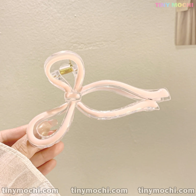 A hand holds Tiny Mochi’s Elegant Bow Hair Clip, a handmade hair accessory featuring a large, translucent bow with pale pink edges. Made from durable plastic for a secure hold. Neutral background with tinymochi.com at the bottom.