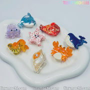 Eight colorful sea creature hair clips from Tiny Mochi are arranged on a white, wavy tray, showcasing these charming mini ocean-themed acetate hair claws for women and girls. The “TINY MOCHI” logo adds a playful finishing touch.