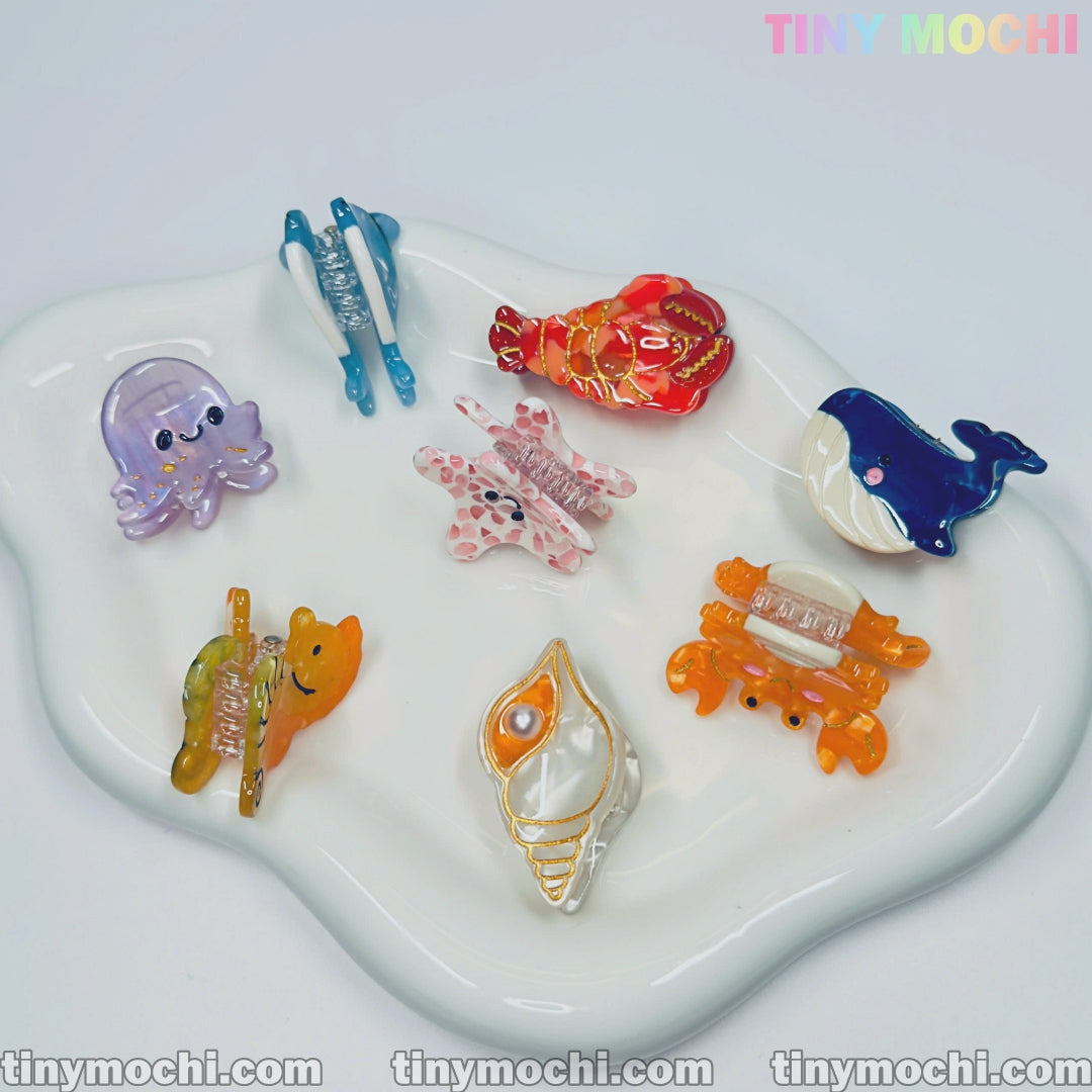A white dish displays eight colorful sea creature hair clips—octopus, lobster, whale, clam, crab, bear, and two bunnies. The text Tiny Mochi appears in pastel colors at the top right. Cute Sea Creature Hair Clips by Tiny Mochi.