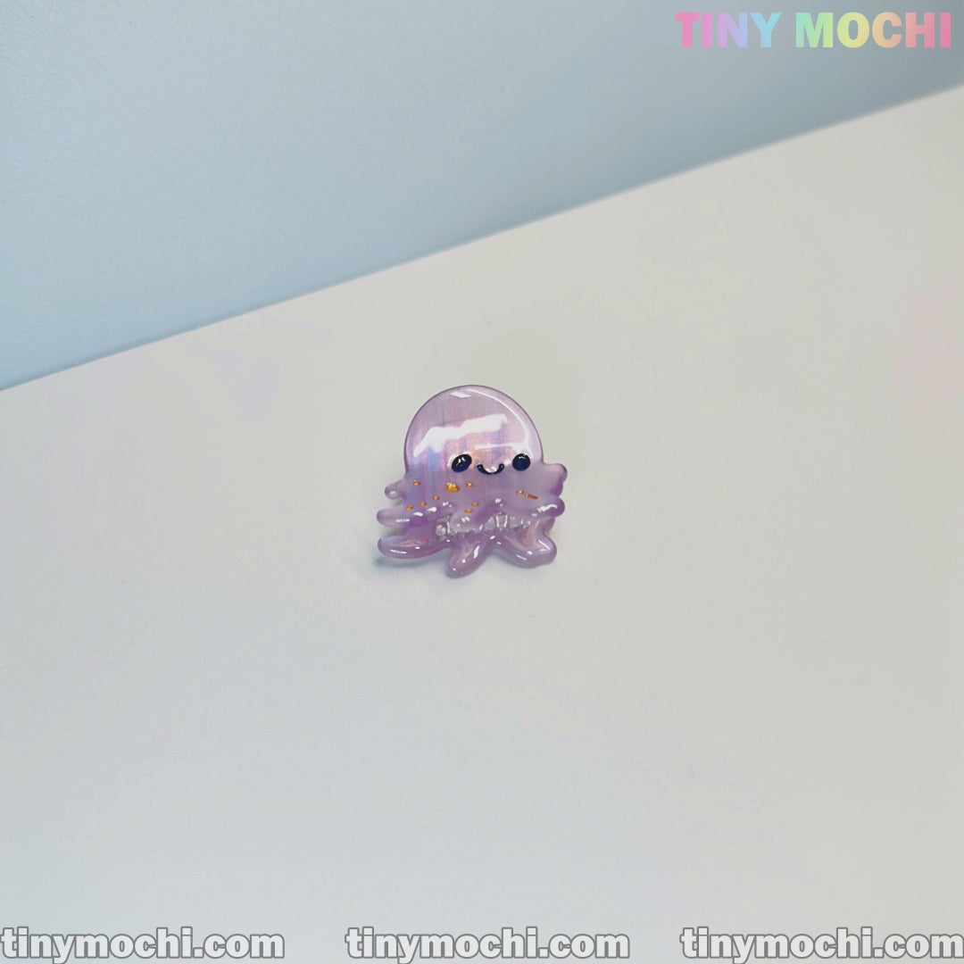A small, shiny purple octopus pin with a cute smile rests on a white surface. The light blue background features Tiny Mochi in pastel—perfect for fans of Cute Sea Creature Hair Clips and ocean-themed accessories for women & girls.