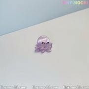 A small, shiny purple octopus pin with a cute smile rests on a white surface. The light blue background features Tiny Mochi in pastel—perfect for fans of Cute Sea Creature Hair Clips and ocean-themed accessories for women & girls.