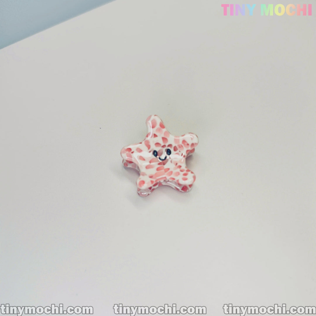 A small, pink and white star-shaped hair clip with a smiling face sits on a white surface. The top right shows colorful Tiny Mochi branding, with tinymochi.com along the bottom—ideal for fans of Cute Sea Creature Hair Clips by Tiny Mochi.