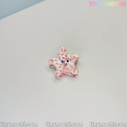 A small, pink and white star-shaped hair clip with a smiling face sits on a white surface. The top right shows colorful Tiny Mochi branding, with tinymochi.com along the bottom—ideal for fans of Cute Sea Creature Hair Clips by Tiny Mochi.