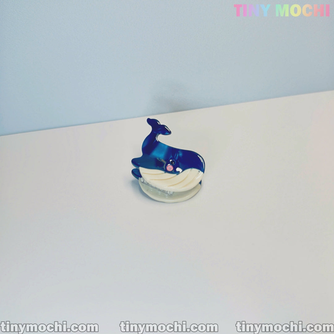 A small, blue and white whale figurine with a smiling face sits on a white surface. Tiny Mochi’s Cute Sea Creature Hair Clips—mini ocean-themed animal acetate hair claws for women & girls—are available now at tinymochi.com.
