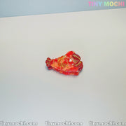 A small, shiny, red and gold hair clip shaped like a sea creature rests on a white surface. The image shows tinymochi.com and Tiny Mochi in pastel at the top right. Product: Cute Sea Creature Hair Clips by Tiny Mochi.