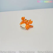 A small, orange crab figurine with big eyes sits on a white surface, holding a seashell, showcasing Tiny Mochi’s Cute Sea Creature Hair Clips—mini ocean-themed animal acetate hair claws for women and girls. TINY MOCHI appears in pastel at the top right.