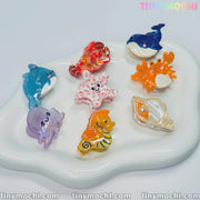 A white dish showcases eight pastel Cute Sea Creature Hair Clips by Tiny Mochi, featuring mini ocean-themed acetate hair claws shaped like a dolphin, whale, octopus, seahorse, crab, shell, and starfish.
