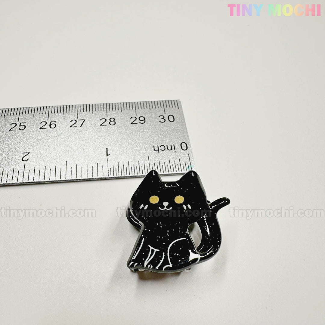 A small black cat enamel pin with yellow eyes sits beside a silver ruler. Pair it with Kawaii Halloween Hair Claw Clips – Set of 8 Spooky-Cute Designs from Tiny Mochi Gift Shop Houston for a cute, standout accessory collection.