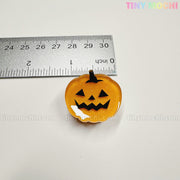 A small orange pumpkin charm with a black jack-o-lantern face, about 1 inch wide, sits below a ruler. Featured as part of Tiny Mochi Gift Shop Houston's Kawaii Halloween Hair Claw Clips – Set of 8 Spooky-Cute Designs.