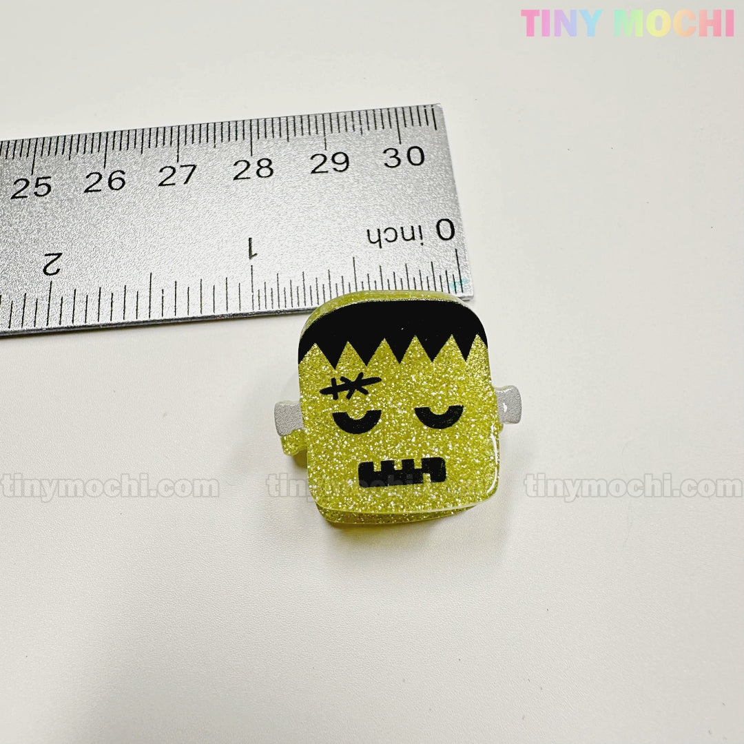 A small glittery yellow Frankenstein's monster from the Kawaii Halloween Hair Claw Clips – Set of 8 Spooky-Cute Designs is shown next to a ruler (about 3 cm tall). Tiny Mochi Gift Shop Houston branding appears in pastel in the corner.